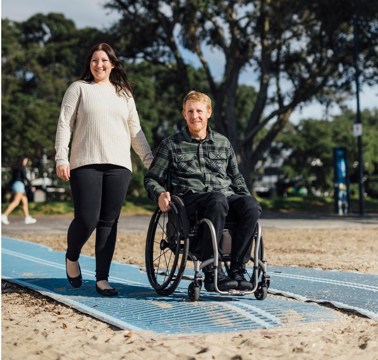 A man on the beach using a wheelchair to move with a woman accompanying him. 