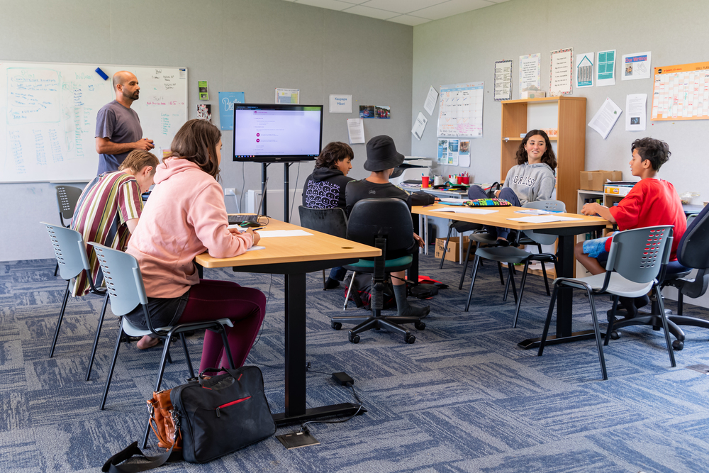 High school age students sitting around two tables in a classroom with a teacher standing by a whiteboard.