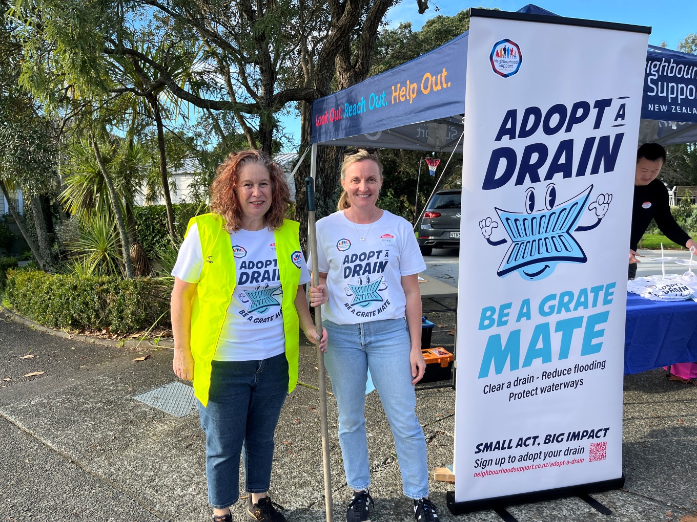 Two women wearing "Adopt A Drain" tshirts next to an Adopt A Drain sign