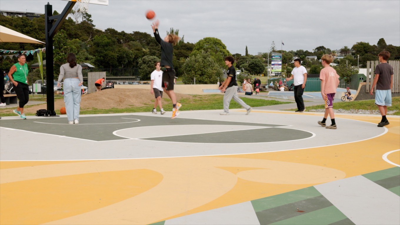 Youth playing basketball on a court in the foreground, with families at a skate bowl and trees in the background.